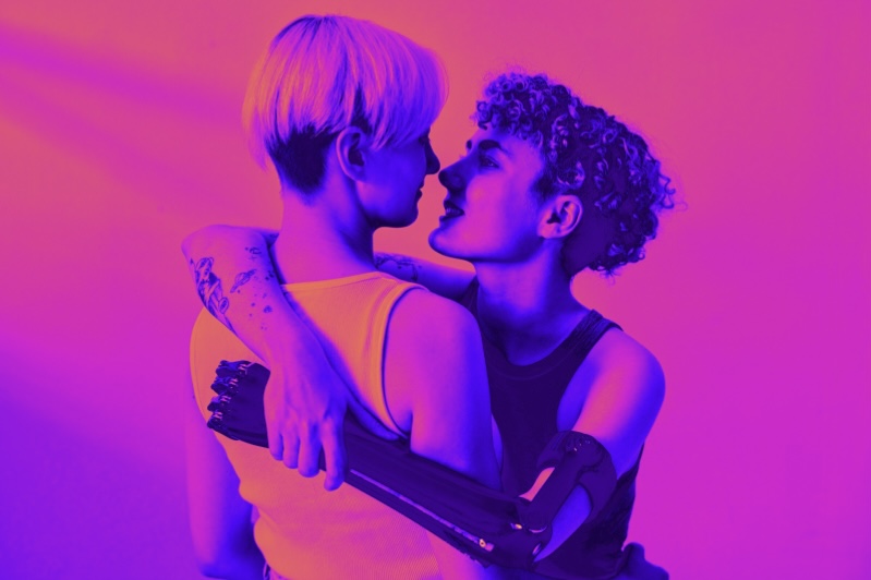 Two people embrace, gazing into each other's eyes.  One has a prosthetic arm and they are both wearing a vest top, looking sexy af against a vivid pink and purple backdrop.