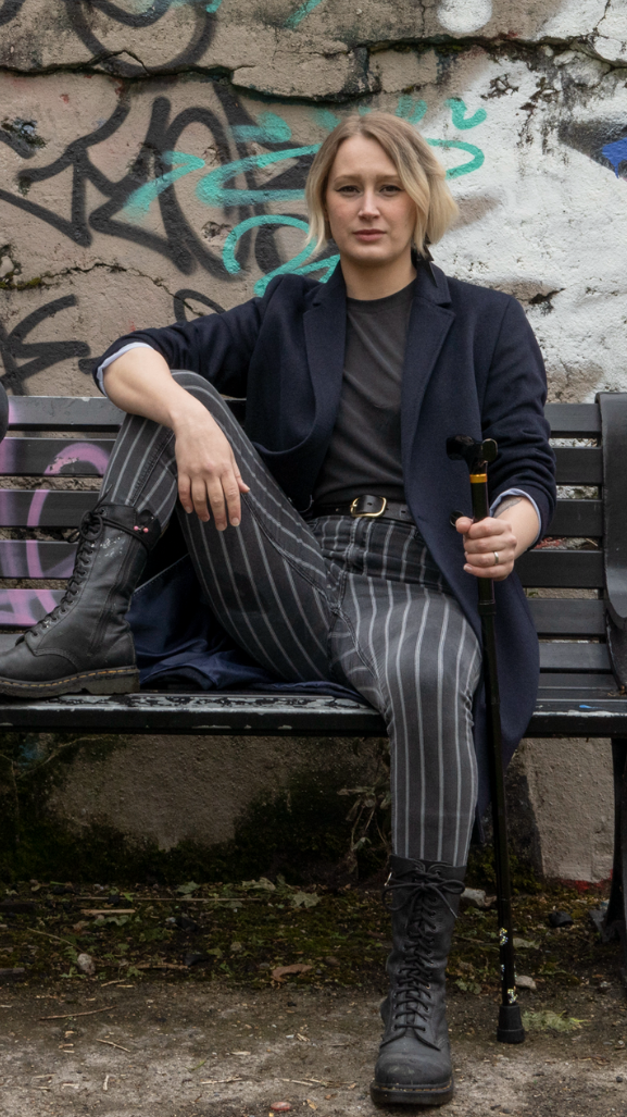 A white person sitting on an outside bench wearing black and white striped trousers, a black top and coat and black boots. They are looking directly at the camera and holding a walking stick in their left hand.